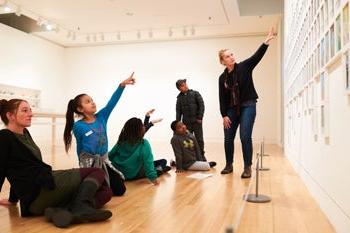 Students point to an art work with a teacher in a gallery