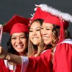 三名学生戴着毕业帽，穿着毕业服，微笑着拍照，中间的一个拿着手机自拍.