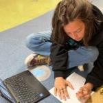 A student sits on a classroom floor drawing on a piece of paper with an open laptop